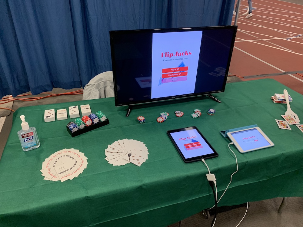 Picture of a table with two iPads, one with Flip Jacks the game on it and another with a newsletter sign up form. A TV is behind the iPads with the iPad showing Flip Jacks on the screen. There are also a bunch of playing cards and poker chips on the table.